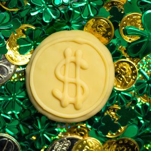 Close-up of a chocolate coin with a dollar sign on a background of green clover leaves and gold coins.